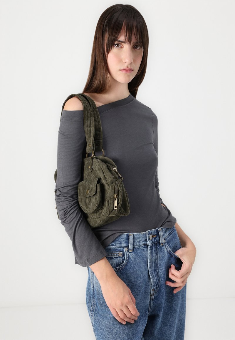 Grauer langärmeliger Oberteil mit einem schulterfreien Design, kombiniert mit hochtaillierten blauen Jeans. Olivgrüne Cordtasche mit Taschen.