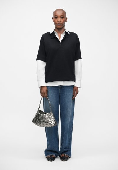 Polo noir à manches courtes sur une chemise blanche à manches longues, associé à un jean bleu à jambes larges et un sac à main en cuir tissé argenté.