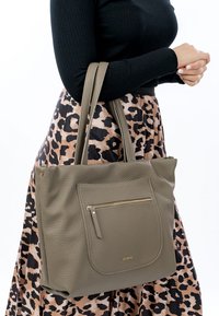 Olive green leather tote bag with textured surface, featuring a front zip pocket and dual handles. Accented with a gold logo.