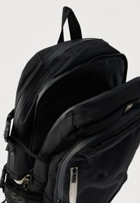 Mochila preta com design estruturado, apresentando um exterior texturizado, compartimentos com fecho de correr e uma pega superior para transporte.