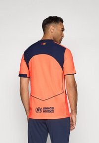 Fényes narancssárga sportmez sötétkék hátrésszel, texturált anyaggal és logóval az alján. Rövid ujjú és kerek nyakú.