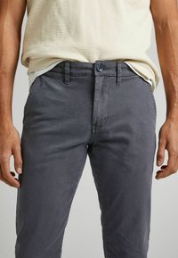 Mężczyzna w ciemnoszarych spodniach i kremowej, strukturalnej koszuli z krótkim rękawem, z rękami swobodnie opadającymi po bokach, sięgającej do talii i ud.