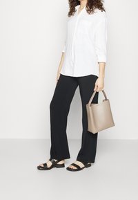 Vit långärmad skjorta med knappar, svarta vida byxor, svarta sandaler och en beige strukturerad tote-väska. Tyget verkar lätt och slätt.