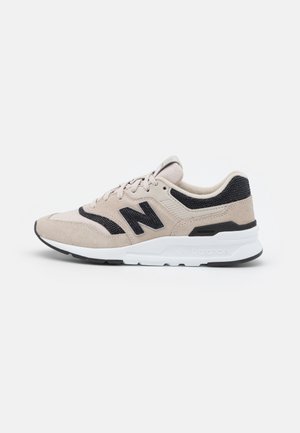 Baskets New Balance beiges et noires avec un design à lacets, semelle rembourrée et logo sur le côté, présentées sur un fond blanc uni.