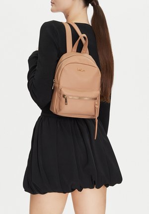 Frau im schwarzen Kleid, die einen hellbraunen DeeZee-Rucksack mit Reißverschlusstasche vorne trägt, von hinten und im Seitenprofil gesehen.