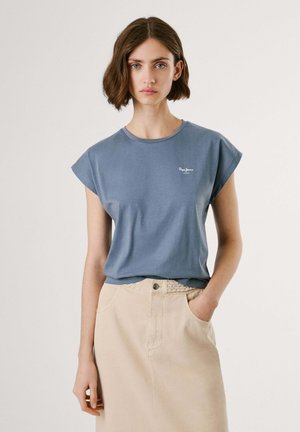 Blue cotton t-shirt with rolled short sleeves and a small white logo on the chest, paired with a light beige skirt.