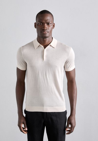 Camisa polo de color beige claro con cuello, mangas cortas y un sutil logo en el pecho. Puños y dobladillo de canalé. Textura suave.