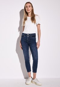 Mörkblå jeans med en figurnära design, utrustade med en knapp- och dragkedjestängning, framfickor och en kort längd. Sammanställda med en vit t-shirt och sneakers.