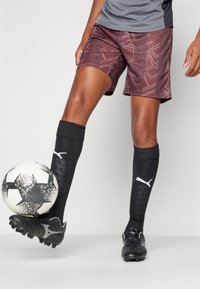 Vinröda shorts med geometriskt mönster, svarta knähöga strumpor och svarta fotbollsskor. En svartvit fotboll är placerad nära foten.