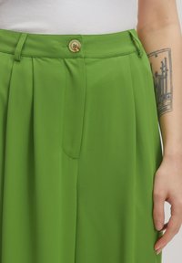 Green pleated trousers with a button closure at the waist, featuring a smooth texture and a side pocket detail.