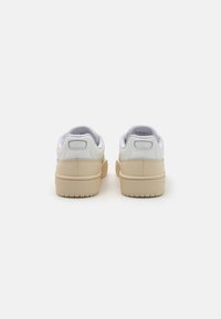 Baskets en cuir blanc avec une semelle en caoutchouc beige, arborant une texture lisse et un col rembourré. La vue arrière montre un branding minimal.
