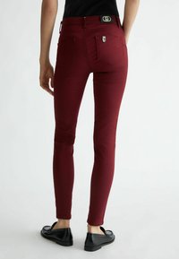 Wąskie jeansy w kolorze bordowym o gładkiej fakturze, z pięcioma kieszeniami i naszywką z logo na pasku. Noszone z czarnymi butami wsuwanymi.