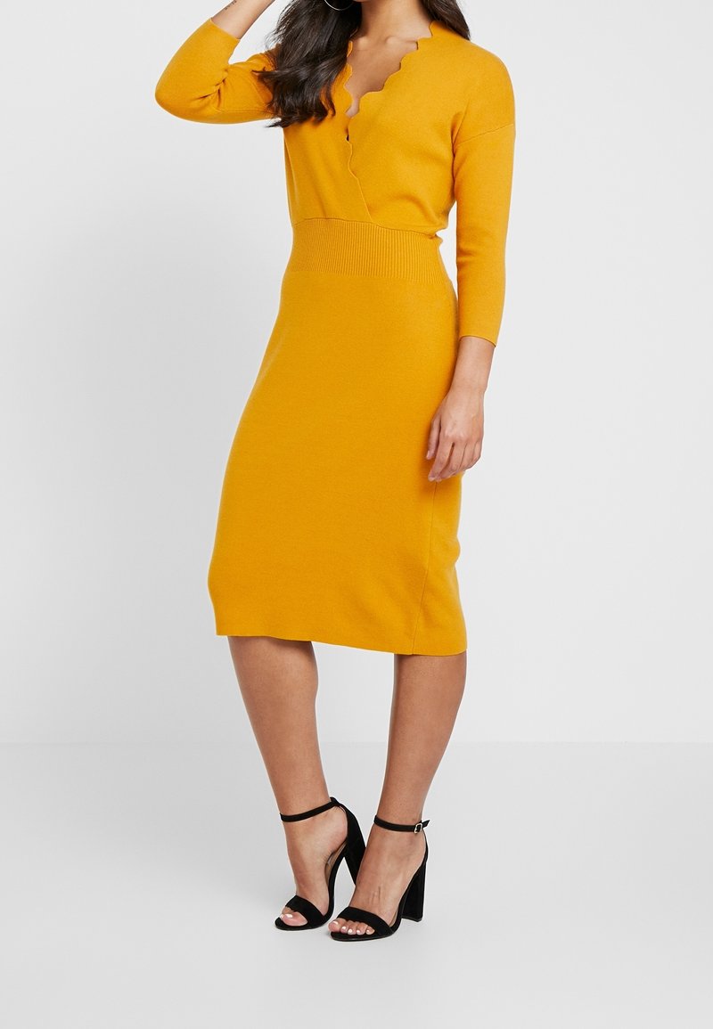 Femme portant une robe jaune moutarde longueur genou avec un col festonné et des manches trois-quarts, associée à des sandales noires à talons hauts.