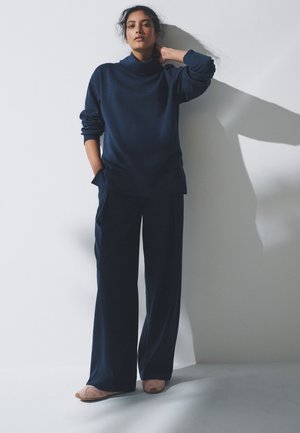 Kobieta w luźnym granatowym swetrze z golfem i spodniach o szerokich nogawkach, stojąca z jedną ręką w kieszeni, a drugą za karkiem, na tle białej ściany.