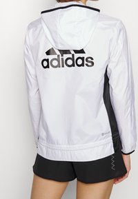 Bílá větrovka Adidas s černými doplňky, velkým černým logem na zádech, lehký materiál a kapuce.