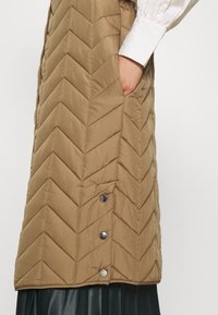 Hnědá prošívaná vesta s chevronovým vzorem, boční kapsou a akcenty na cvoky; má lesklou texturu látky a vrstvenou černou sukni.