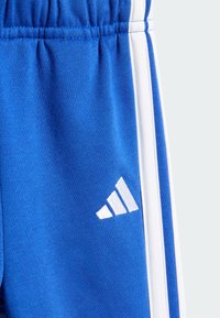 Pantalons de sport bleus en matériau doux, avec une texture lisse, des bandes latérales blanches et un accent de logo Adidas blanc.