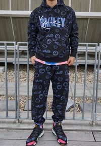 Osoba v černé mikině s kapucí a kalhotách s modrými usmívajícími se tvářičkami a textem, stojící u kovového plotu na kamenném a dřevěném povrchu.