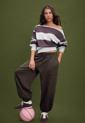 Felpa a righe oversized grigia e marrone, abbinata a joggers larghi color oliva scuro. Il modello è in piedi su un pallone da basket rosa contro uno sfondo verde.