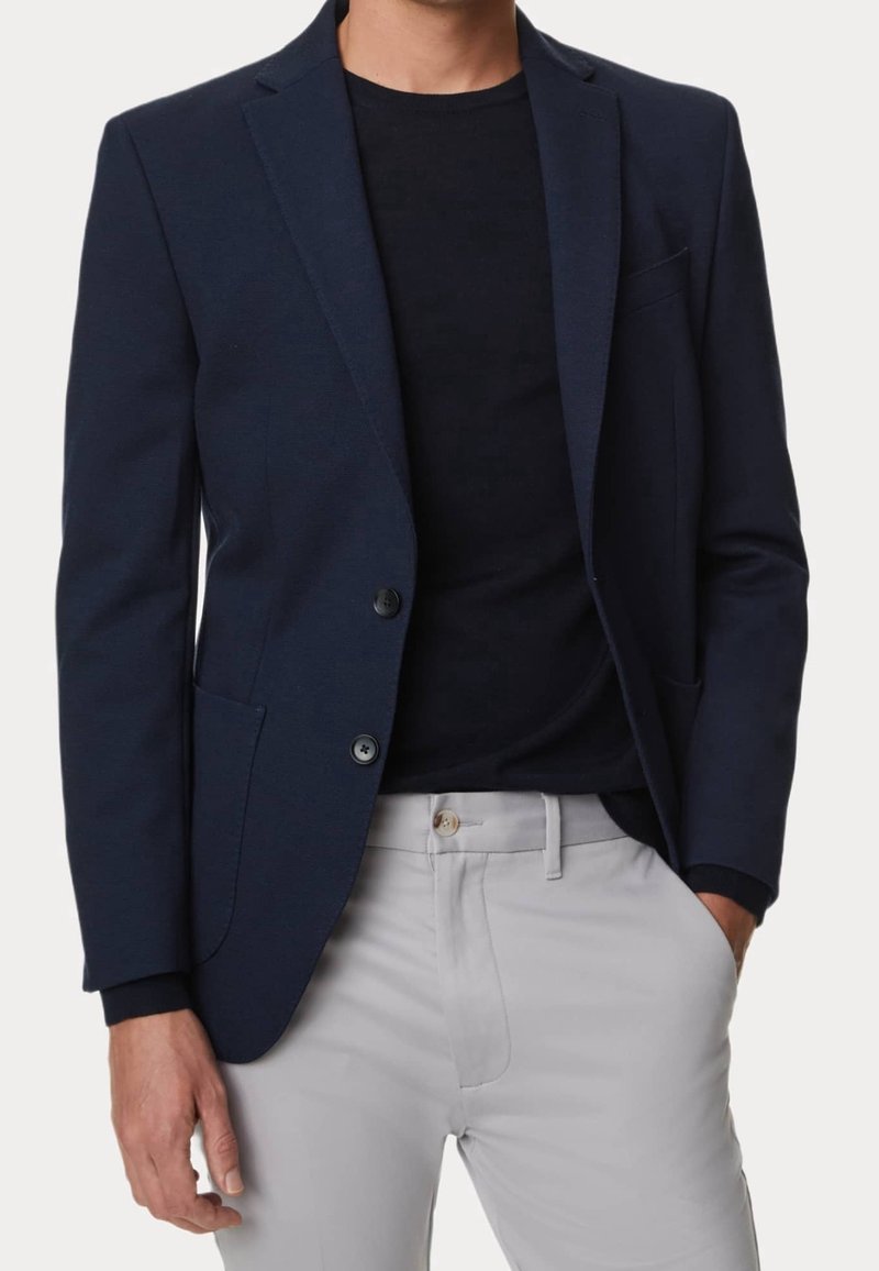 Blazer navy con due bottoni, rever a scendere e tessuto testurizzato, abbinato a una camicia nera e pantaloni grigio chiaro.