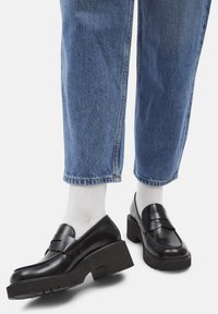 Persona con calcetines blancos, mocasines de plataforma gruesa negros y jeans de mezclilla azules frente a un fondo claro y liso.