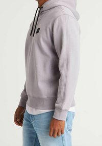 Light gray hoodie with a drawstring hood, black logo on the chest, ribbed cuffs, and waistband, displayed on a person. Casual fit.