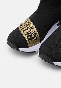 Černé tenisky v ponožkovém stylu mají texturovaný svršek se zlatým logem s nápisem "VERSACE JEANS COUTURE", bílé mezipodešve a černé podešve.