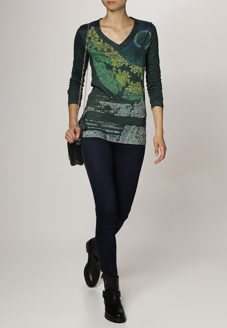 Haut vert à manches longues et décolleté en V profond, avec des motifs tourbillonnants multicolores complexes, associé à un jean skinny noir et des bottines noires.