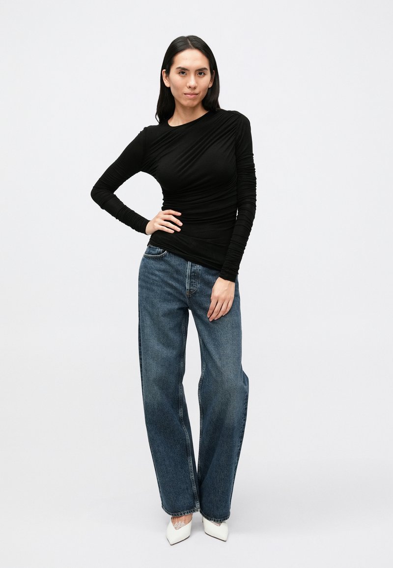 Sort langærmet top med draperet design, kombineret med lyseblå vidde-ben jeans og hvide spidse sko. Ensfarvet baggrund.