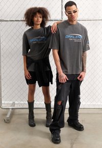 Grå oversized t-shirts med "OFFLINE FOR PRIVATE REASONS" i blå, parret med slidte sorte jeans og knæhøje sorte støvler. Hvide solbriller bæres.
