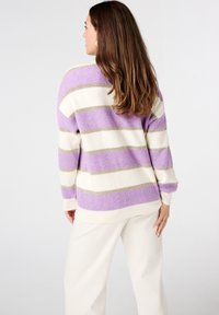 Gestreepte trui in lavendel en crème met gouden accenten. Ribbeltextuur, ruime mouwen en een relaxed fit. Gecombineerd met lichtgekleurde spijkerbroek.