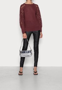 Burgunderfarbener Strickpullover mit Spitzenärmeln, kombiniert mit schwarzen Lederleggings und einer silbernen metallischen Handtasche mit Kettendetails und einem kurzen Griff.