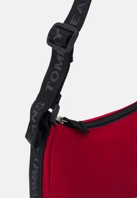 Bolsa de tela roja con un acabado texturizado, que cuenta con una correa negra ajustable con un logotipo y un cierre con cremallera.