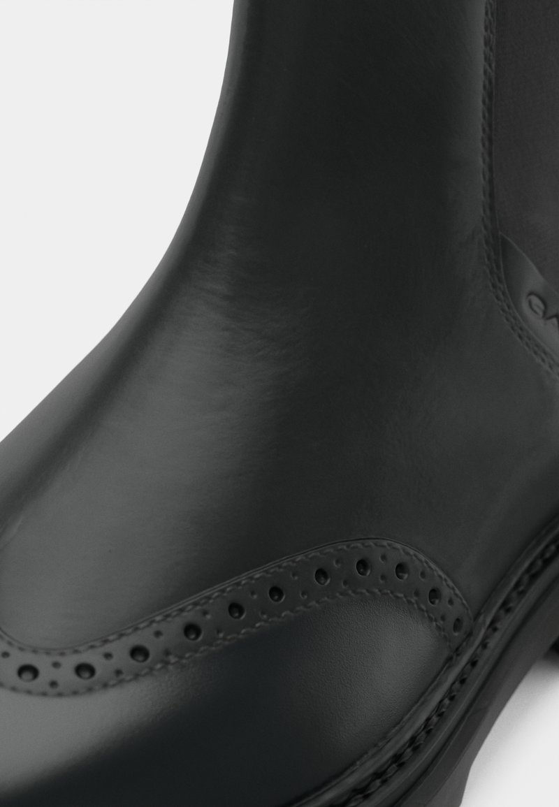 Schwarzer Gummistiefel mit einer glatten Oberfläche, runder Form, Broguemuster an der Seite und einer strukturierten Sohle für besseren Grip.