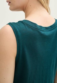 Top fără mâneci, de culoare teal, cu guler rotund, realizat dintr-un material moale. Textură netedă, având cusături minime pe cusăturile umerilor.