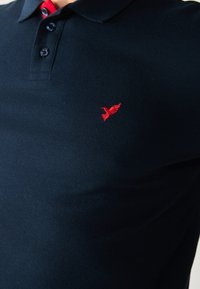 Polo bleu marine avec un design collier, trois boutons et un logo d'oiseau rouge brodé sur le côté gauche de la poitrine. Tissu texturé.