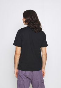 T-shirt en coton noir à manches courtes, présentant un col arrondi classique et un dos uni. Porté avec un pantalon cargo violet.