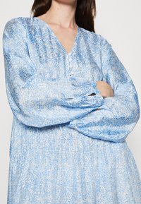 Vestido azul claro con un estampado floral, que presenta un escote en V, detalles de botones y puños fruncidos. Está hecho de una tela fluida, con un corte holgado.