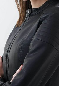 Persona che indossa una giacca di pelle nera con texture, pannelli imbottiti sulle spalle, bottone a scatto sul colletto e cerniera frontale, con le braccia incrociate.