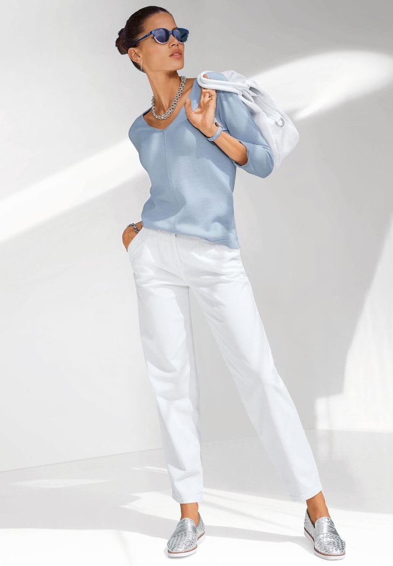 Lichtblauw, langemouwen top met een V-hals; witte broek; zilveren instap schoenen met een textuurpatroon; een witte tas; blauwe zonnebril.