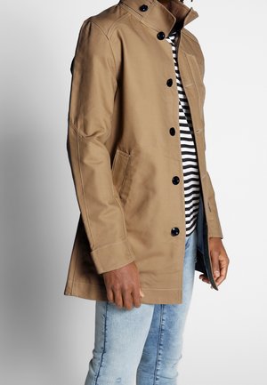 Homme portant un manteau boutonné couleur fauve sur une chemise rayée noir et blanc et un jean bleu clair, debout devant un fond uni.