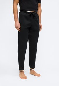 Άνδρας με μαύρο παντελόνι φόρμας με κορδόνι στη μέση και ριγωτές μανσέτες, όρθιος ξυπόλητος σε απλό λευκό φόντο.