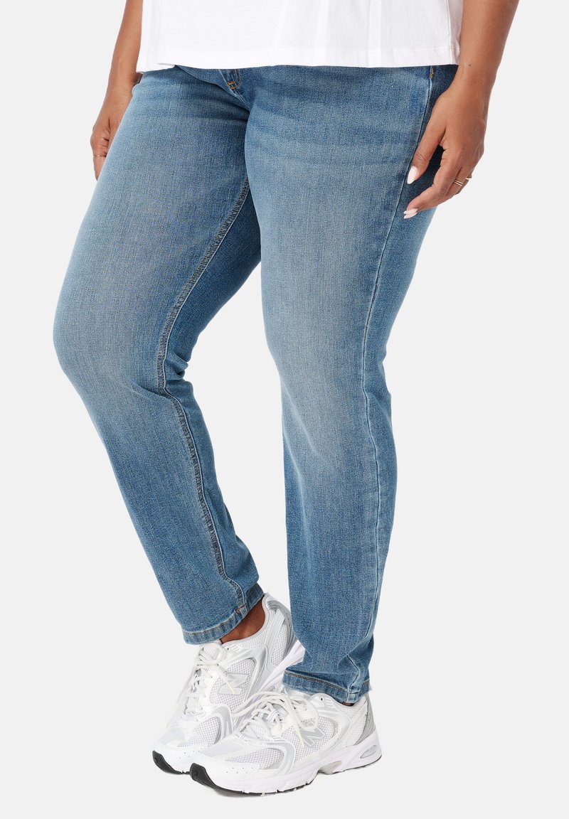 Persoon in lichtblauwe jeans en witte sneakers, staande met ontspannen handen langs de zij op een witte achtergrond.