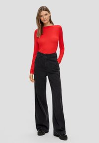 Rotes Langarmshirt kombiniert mit hoch taillierten schwarzen weit geschnittenen Hosen, mit glatter Textur und klassischem Knopfverschluss. Schwarze Plateauschuhe.
