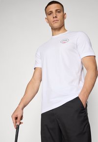 Weißes kurzärmliges T-Shirt mit rundem Ausschnitt und einem kleinen Blumenlogo-Patch auf der Brust, kombiniert mit schwarzen Hosen. Einfaches Design.