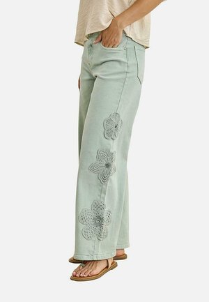 Mujer con vaqueros de pierna ancha de color verde claro con tres grandes aplicaciones de encaje floral en la pierna izquierda, combinados con top beige y sandalias.