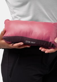 Cuscino portatile rosa con una texture liscia e una cerniera viola scuro alla base, etichettato "STOW AWAY" in bianco. Sostenuto da due mani.