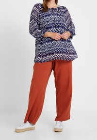 Langærmet, marineblå bluse med et geometrisk hvidt mønster og en bindebånds-halsudskæring, parret med løse, terrakottafarvede bukser og hvide sneakers.