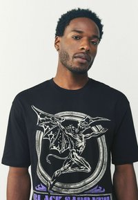 Mand med sort t-shirt med et stort hvidt og lilla grafisk billede af en vinget dæmon og teksten "Black Sabbath".