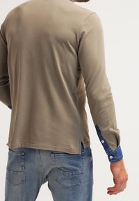 Chemise à manches longues, de couleur olive, avec un ourlet arrondi. Dotée d'une accent en denim sur les poignets et d'une texture douce et lisse.